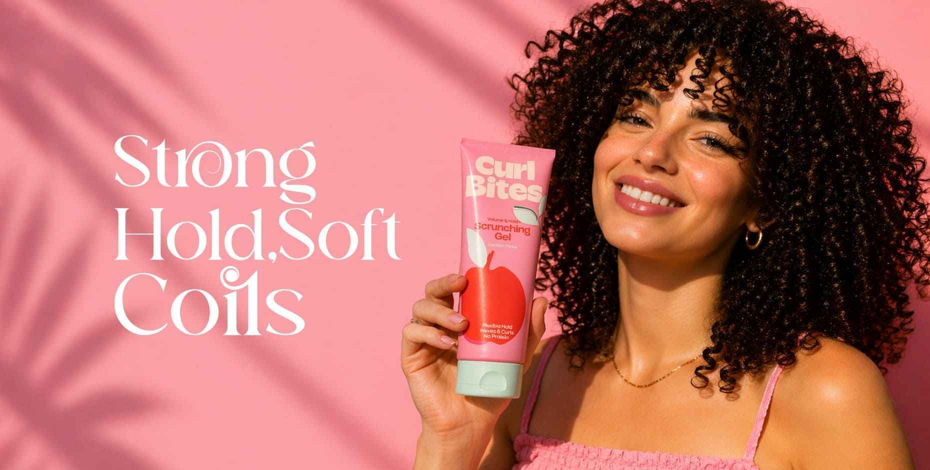 Smiling woman with voluminous coily hair holds a pink tube of Curl Bites Scrunching Gel.