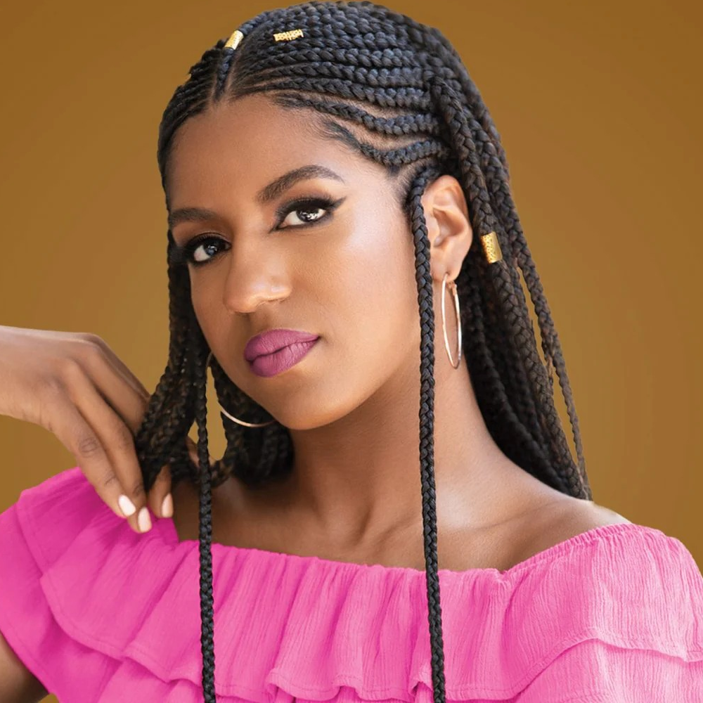 Woman with long black braids, gold cuffs, hoop earrings, pink off-shoulder top, and magenta lipstick.
